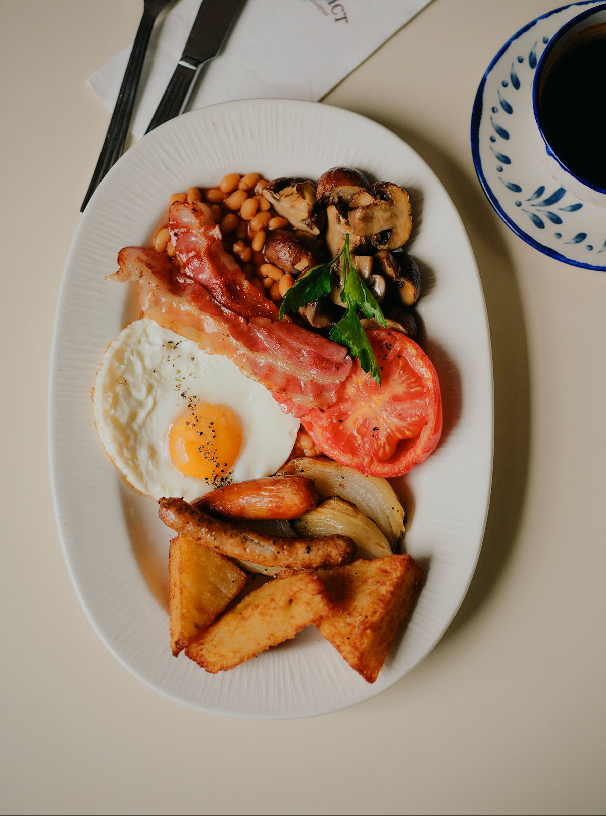 A full English breakfast served on a white oval plate, featuring a sunny-side-up egg, bacon, baked beans, mushrooms, a tomato slice, sausage links, fried potatoes, and a garnish of green leaves. A cup of black coffee on a blue patterned saucer and a fork and knife on a napkin are visible on a beige table.